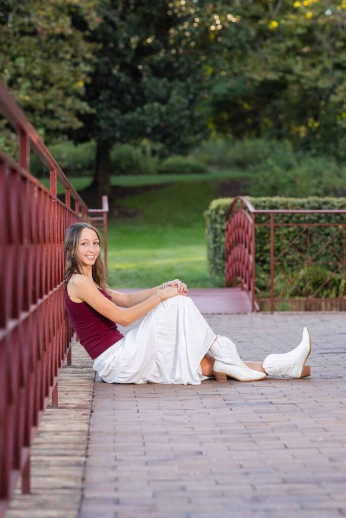 High school senior girl sitting on the ground leaning against a fence in a park